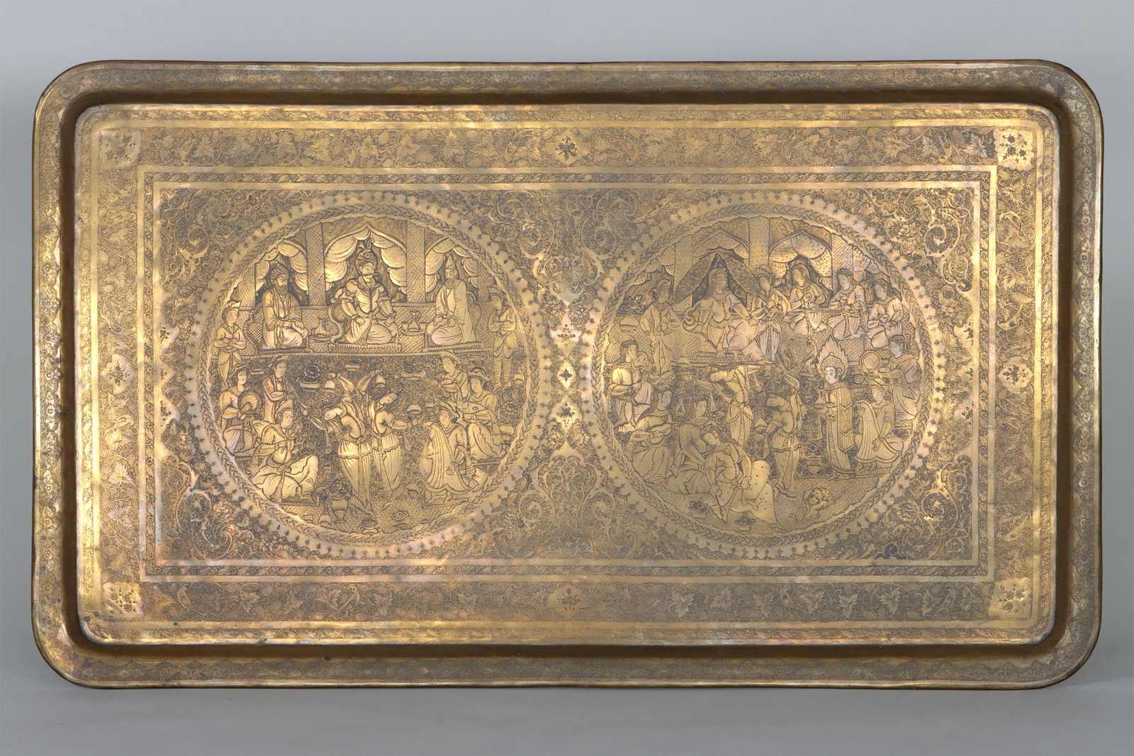 Large Persian Engraved Brass Tray: Large Persian Engraved Brass Tray of rectangular form, with a pair of interior scenes at center depicting dancers and musicians performing for a courtly audience, the scenes enclosed by round scrolled