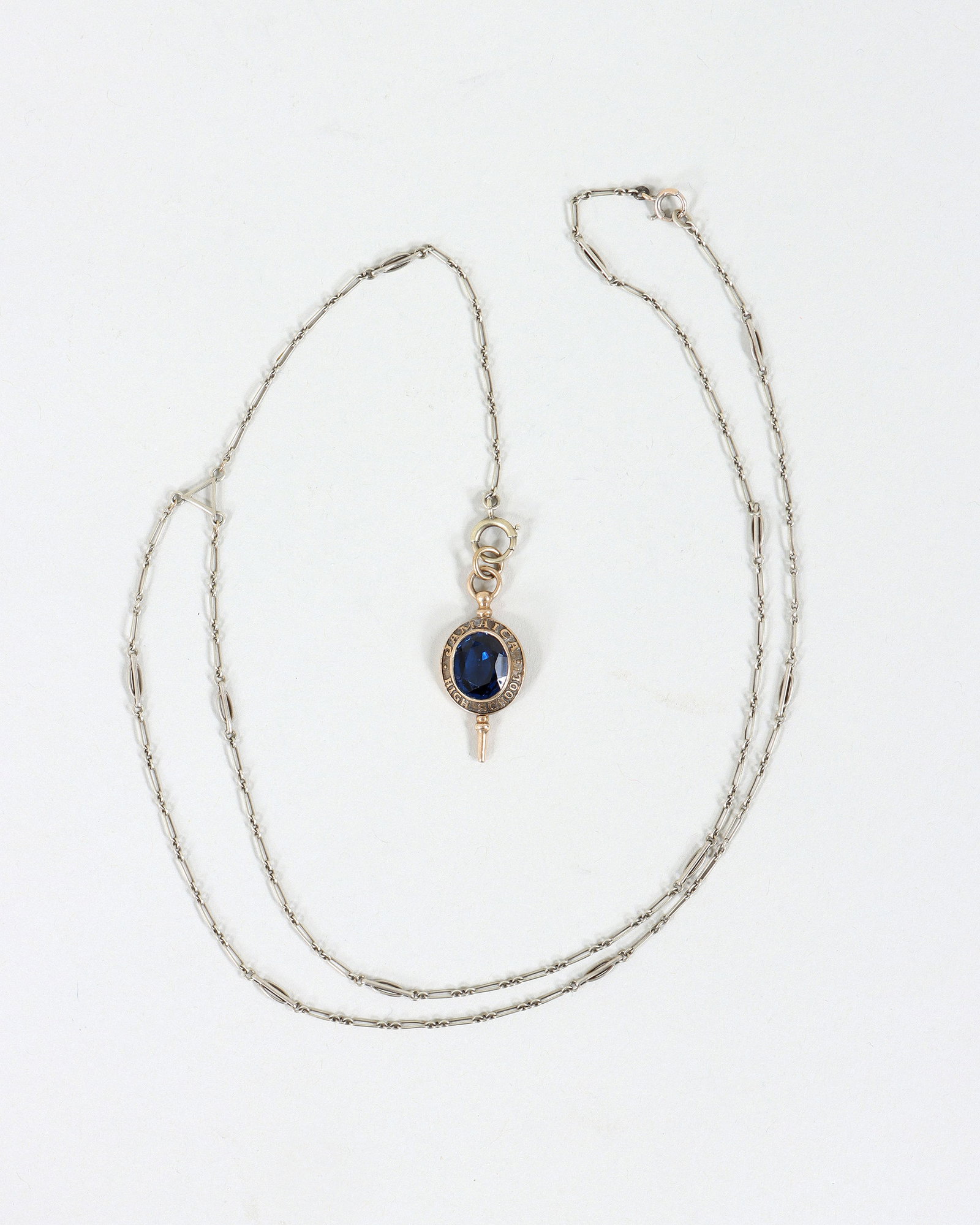 10k Gold Pendant Necklace: 10k Gold Pendant Necklace having Long Island "Jamaica High School" pendant with an oval-cut semi-precious blue stone, back inscribed "L.G. '63", on white gold chain having alternating long and short l