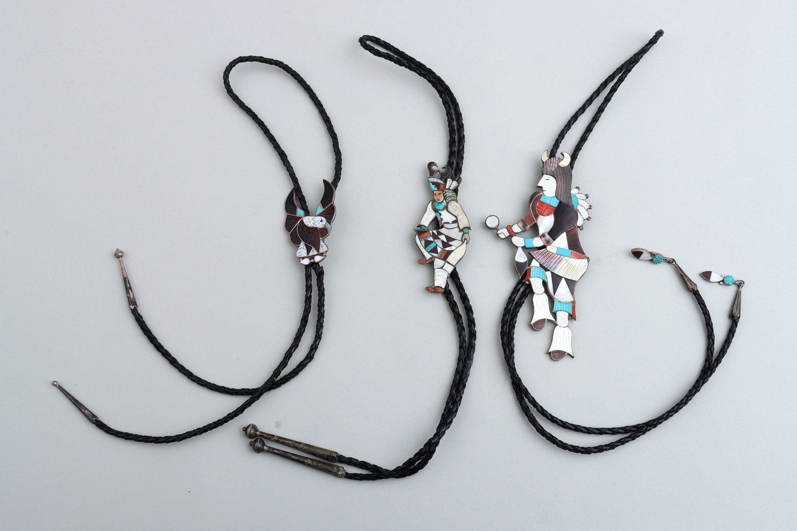 Zuni Carved Mosaic Inlay Silver Bolo Ties: Group of (3) Zuni Carved Mosaic Inlay Silver Bolo Ties all having mosaics with carved decoration inlaid on figural silver bolos, the bolos on braided leather cords ending in silver tips with ball fini