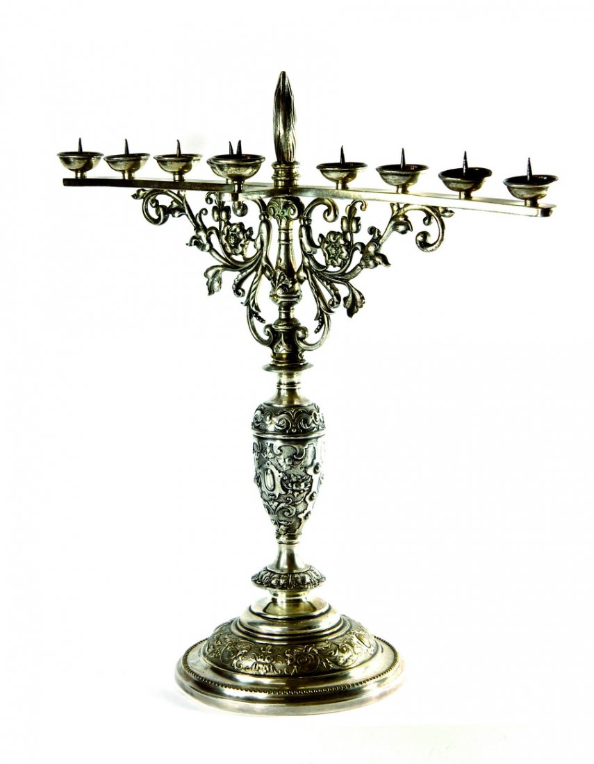 Chanuka Menorah. Germany? First Half of the 20th: Chanuka menorah for candles, casting and boring work. Maker's mark 'W.M.'. Tall menorah, standing on one leg. Wide, rounded base. Floral decorations around the base. Smooth, narrow plank on the top se