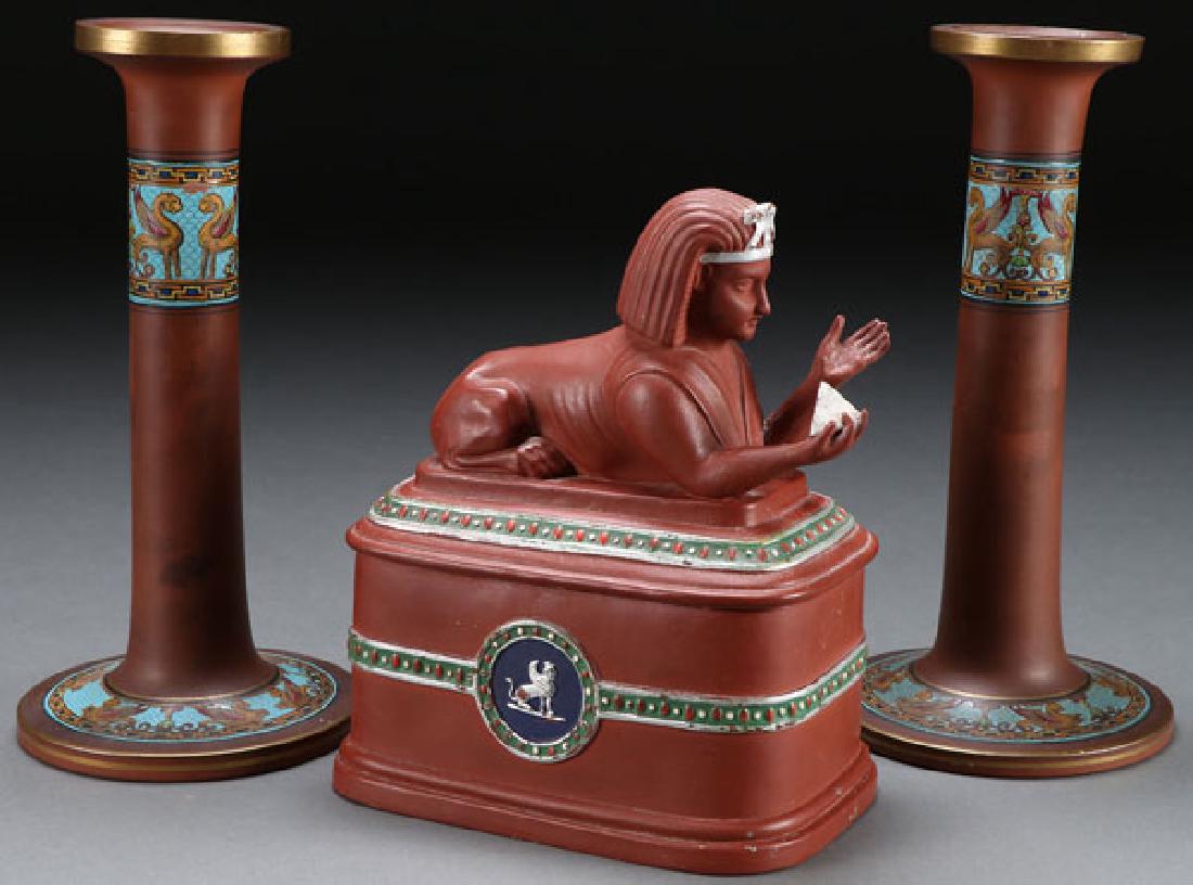 A THREE PIECE GROUP OF EGYPTIAN REVIVAL REDWARE: A THREE PIECE GROUP OF EGYPTIAN REVIVAL REDWARE, 19TH CENTURY. Comprising a pair of candlesticks with enameled sphinx and impressed S on base and a figural covered box with sculpted sphinx lid. The b