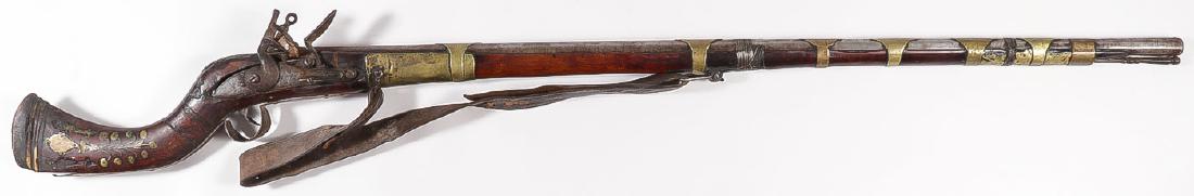 AN INDO-TIBETAN MUSKET: AN INDO-TIBETAN MUSKET. The octagonal twist barrel with flared smooth boar muzzle held in a wood stock with brass barrel bands displaying a flint lock and inlaid stock, leather sling, and metal ra
