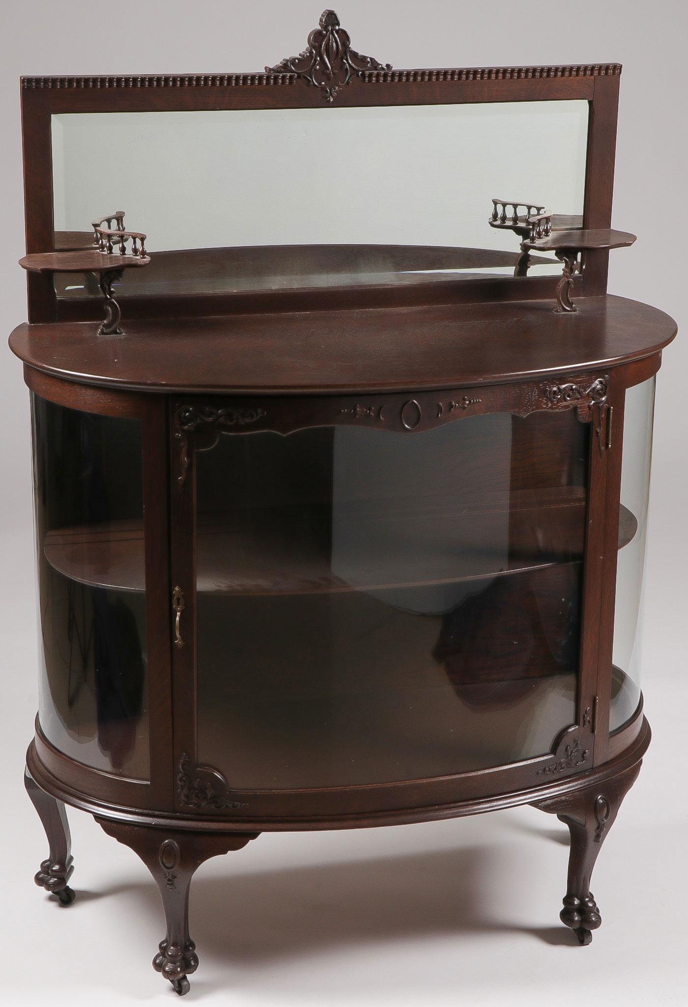 OAK CURVED GLASS MIRRORED BUFFET: OAK CURVED GLASS MIRRORED BUFFET. With a beveled mirror over a demilune molded top resting on conforming case with curved glass sides and central panel door, and raised on short cabriole legs terminat