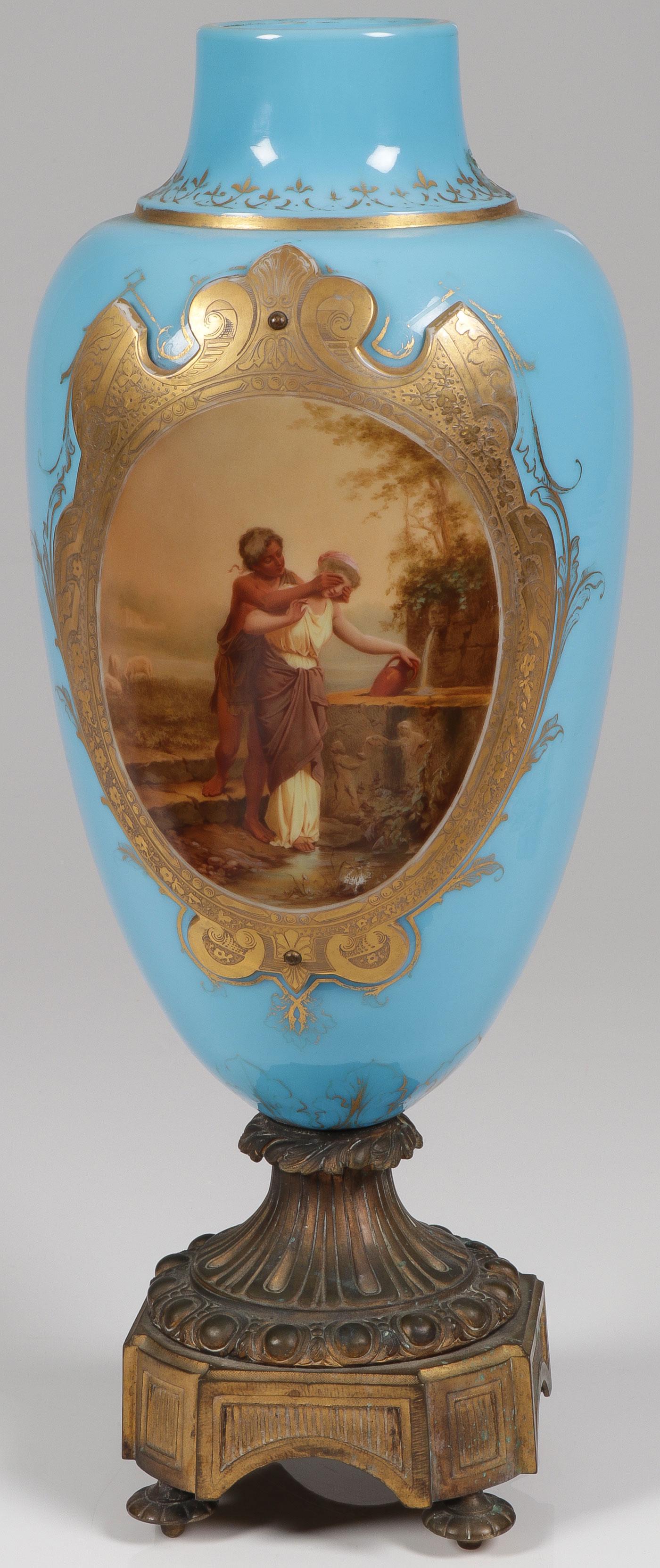 ORMOLU MOUNTED MANTEL VASE: ORMOLU MOUNTED MANTEL VASE. Comprising a blue opaque glass urn formed vase with applied etched glass shield form cartouche with amorous scene at a well, and on a gilt bronze base with bun feet. Height