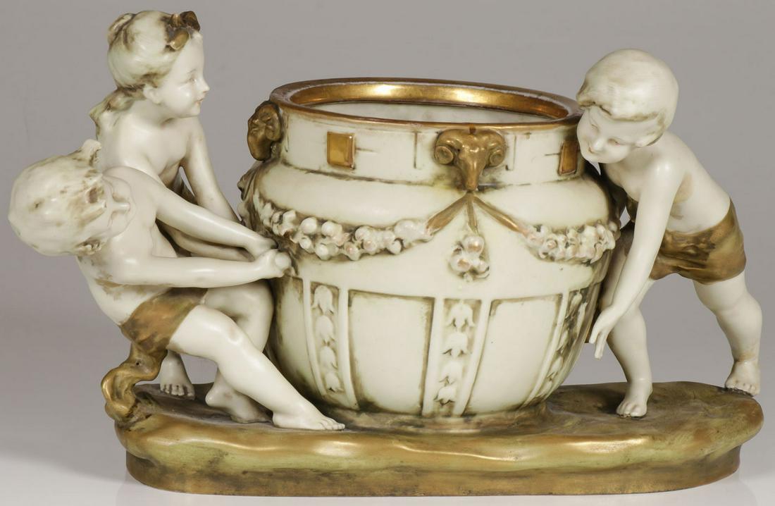 AN AMPHORA PORCELAIN FIGURAL JARDINIÈRE: AN AMPHORA PORCELAIN FIGURAL JARDINIÈRE. Comprising a charming figural grouping of putti moving a jardinière with floral swags and the rim with gilded rams heads, underside with impressed Amphora ma