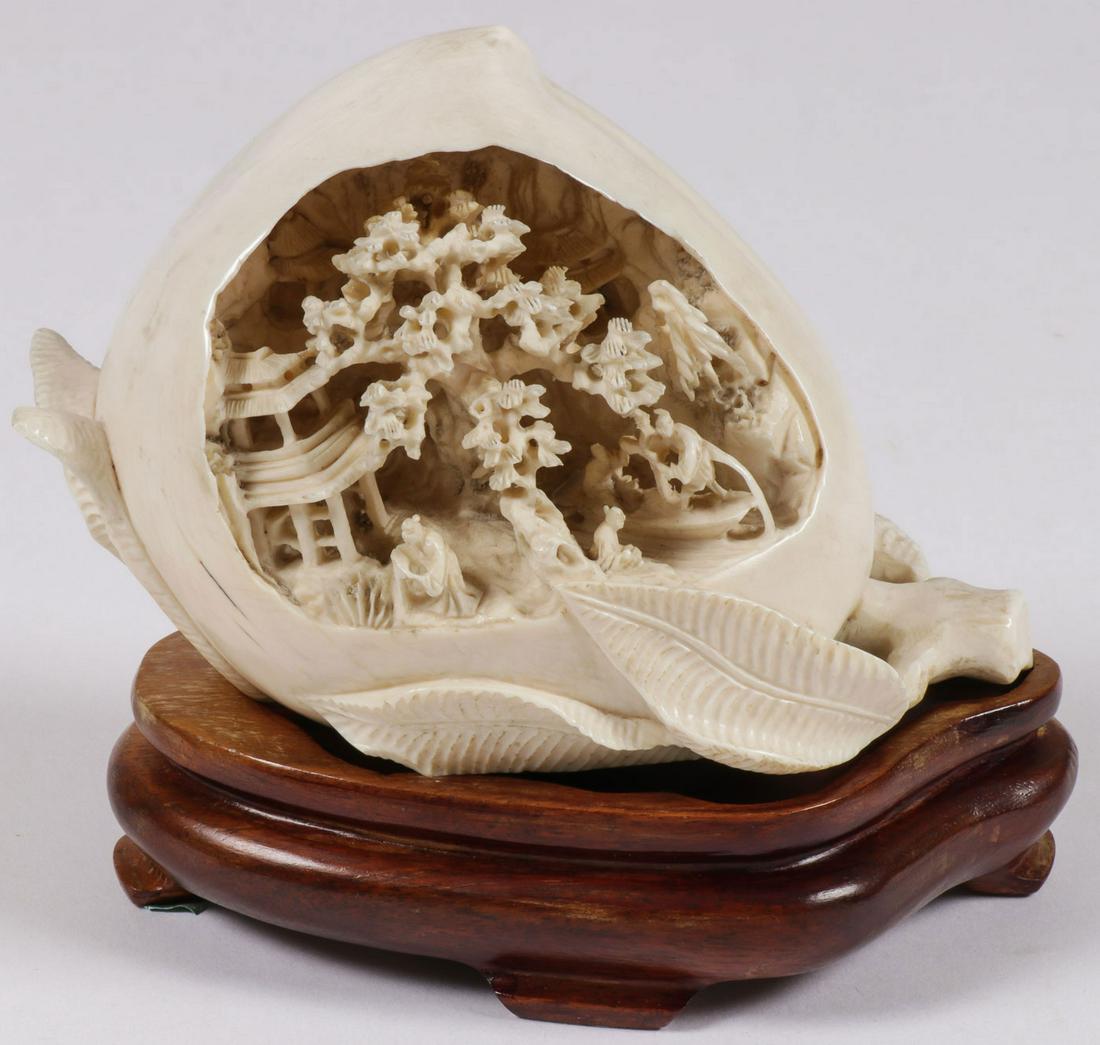 CHINESE CARVED PLUM CIRCA 1900: CHINESE CARVED PLUM CIRCA 1900. Comprising a finely carved plum hollowed out and deeply carved with a village mountain scene in miniature detail. Together with a conforming carved wood base. The carve