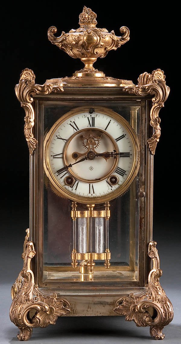 CLOCK: ANSONIA CRYSTAL REGULATOR: AN ANSONIA GILT BRONZE CRYSTAL REGULATOR, circa 1900, with beveled glass panels below an urn shaped finial. Height 15.25 inches (38.7 cm). Estimate $300-$500 Euro 200-400