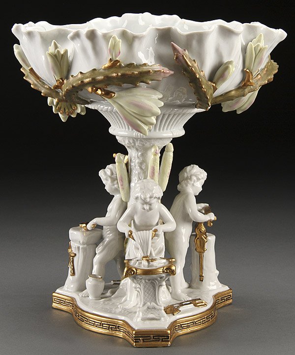 A JAPANESE FIGURAL COMPOTE IN THE EUROPEAN STYLE: A JAPANESE FIGURAL COMPOTE IN THE EUROPEAN STYLE, LAST HALF 20TH CENTURY. The lily form bowl with applied florals above a figural stem with three allegorical putto fashioning cupid's arrows. Height 12