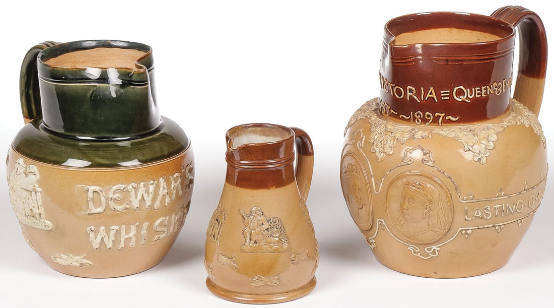 THREE ROYAL DOULTON LAMBETH JUGS: THREE ROYAL DOULTON LAMBETH JUGS. Each with applied designs and handles, including an example commemorating Queen Victoria's reign 1837 ? 1897 and one that reads "DEWAR'S WHISKY," greatest height 6.5 