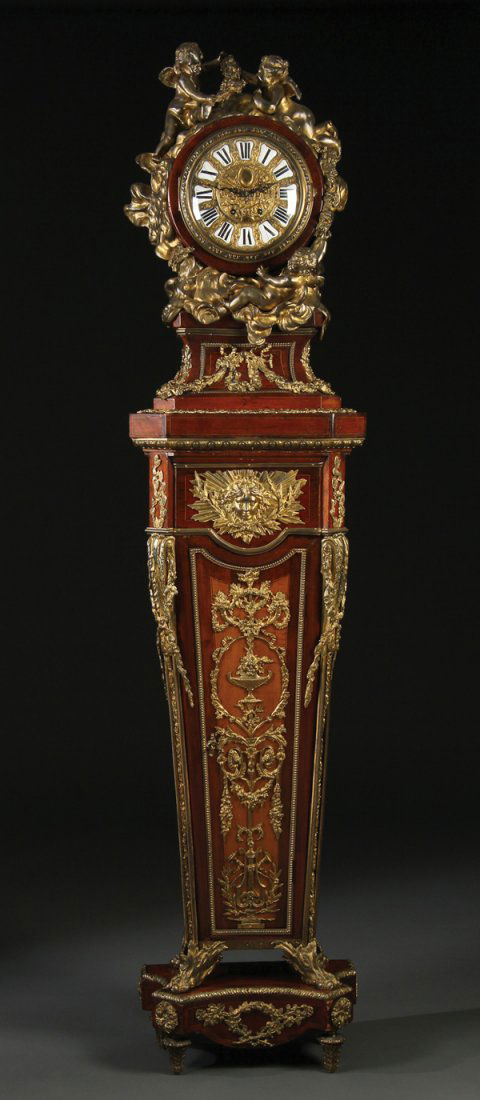 LOUIS XVI STYLE TALL CLOCK, AFTER SORMANI: A GOOD LOUIS XVI STYLE GILT BRONZE AND MAHOGANY LONG CASE CLOCK, AFTER PAUL SORMANI, 19TH CENTURY. The waisted circular clock case mounted with large gilt bronze winged putti resting on bronze cloud