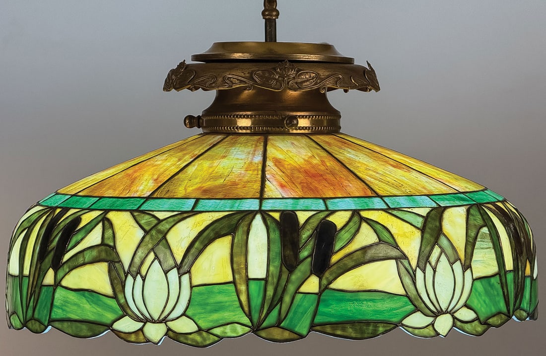 ART NOUVEAU CATAIL & LILY CHANDELIER, C. 1910: ART NOUVEAU CATAIL & LILY CHANDELIER, C. 1910. Comprising a good American leaded glass chandelier, the rim banded in leaded waterlilies and cattails beneath geometric panels, held in a superb gilt bro