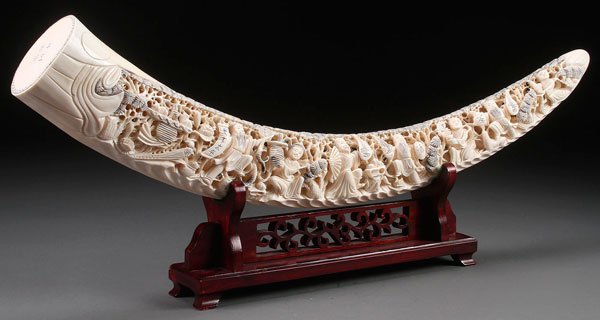 A CHINESE CARVED IVORY TUSK: A CHINESE CARVED IVORY TUSK. Carved in reticulated relief with 14 figures in various activities within flowering pine trees. Displayed on a carved hardwood stand. Length 28 inches (71 cm).