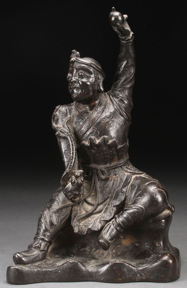 A FINE CHINESE BRONZE FIGURE OF A CUSTODIAN GOD: A FINE CHINESE BRONZE FIGURE OF A CUSTODIAN GOD, QING DYNASTY, PERHAPS EARLIER. Depicting the seated figure with incised robes holding a dragonette in right hand with the left arm raised holding a bal