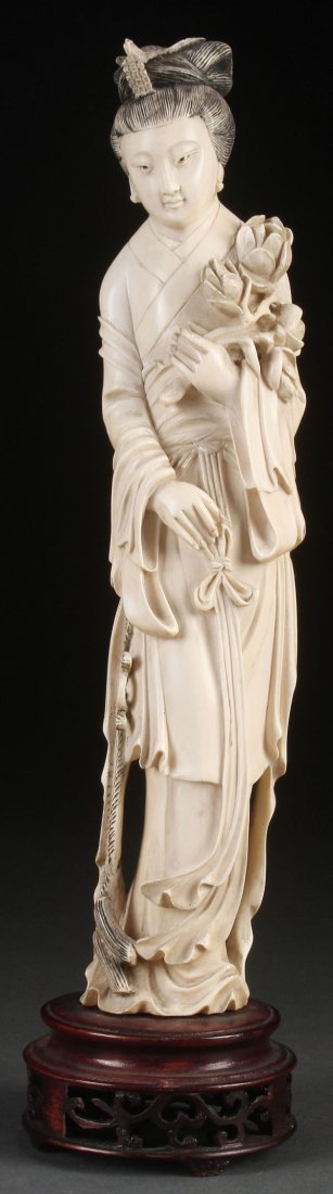 A CHINESE CARVED IVORY KWAN YIN, QING DYNASTY.: A CHINESE CARVED IVORY KWAN YIN, QING DYNASTY. Carved in standing form with flowing robes, holding a lotus blossom. Raised on a pierced carved hardwood stand. Height of ivory 11.75 inches (30 cm). Ove