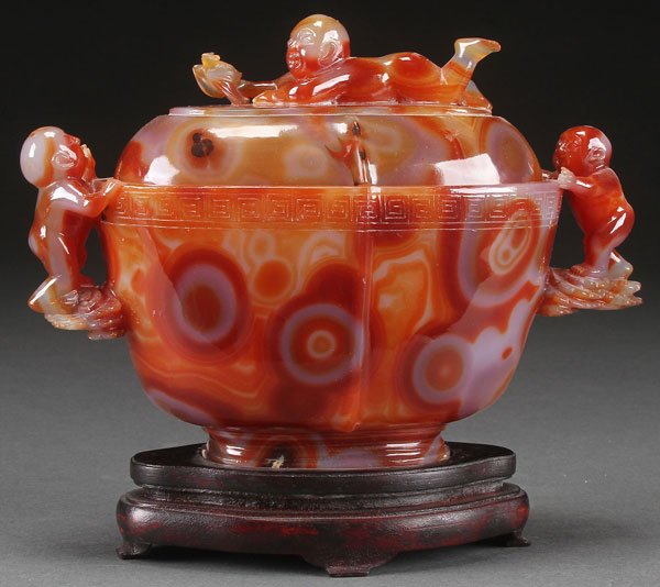 A CHINESE CARVED CARNELIAN AGATE COVERED COUPE: A CHINESE CARVED CARNELIAN AGATE COVERED COUPE. With figural handles and domed lid surmounted with a boy catching a bat. Raised on a fitted hardwood stand. Length 9 inches (23 cm).