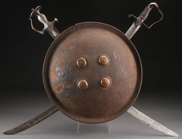 VINTAGE PAIR OF CROSS SWORDS AND SHIELD: A DECORATIVE VINTAGE PAIR OF CROSS SWORDS AND SHIELD. Comprising a Middle Eastern sabre and a British sabre mounted beneath a domed pressed leather (?) shield.