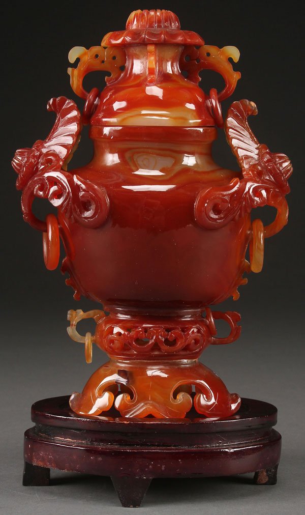 A CHINESE CARVED CARNELIAN AGATE LIDDED URN: A CHINESE CARVED CARNELIAN AGATE LIDDED URN, LAST HALF OF THE 20TH CENTURY. Carved and pierced with foliate form handles, archaic scroll work, and suspended loose rings, raised on a hardwood stand. Ag
