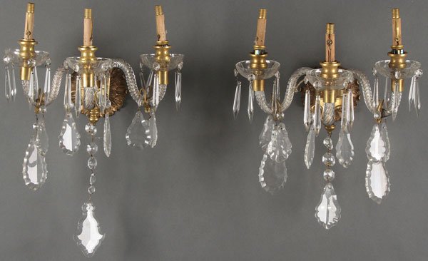 FRENCH GILT BRONZE CUT CRYSTAL WALL SCONCES: A PAIR OF GILT BRONZE AND CRYSTAL WALL CANDELABRA, EARLY 20TH CENTURY. Each with three scrolled twist arm glass supports and with suspended cut glass prisms and pendants, mounted on a sculpted acanthu