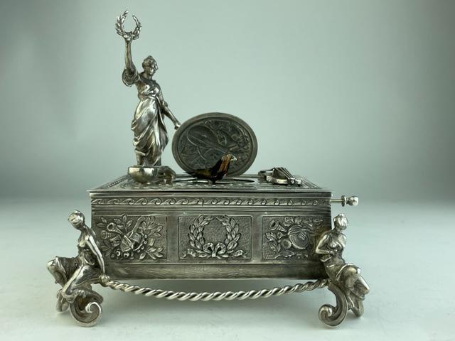 Mechanical singing bird box decorated with a lady: Mechanical singing bird box decorated with a lady holding a wreath in he hand over her head. Each corner supported with a scrolled foot and semi-nude woman. When the knob is pulled the lid raises and 