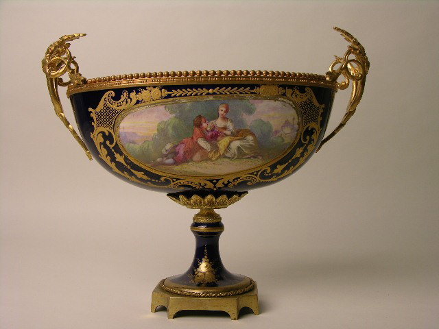 SEVRES CENTERPIECE BOWL WITH GOLD OMULO HANDLES.: A NICE SEVRES CENTERPIECE BOWL WITH GOLD OMULO HANDLES. TWO PAINTED CARTOCES ON SIDES, ONE WITH A RIVER SCENE THE OTHER OF A COURTSHIP. INSIDE THE BOWL PAINTED SPRAYS OF FLOWERS. OVERALL HEIGHT FROM B