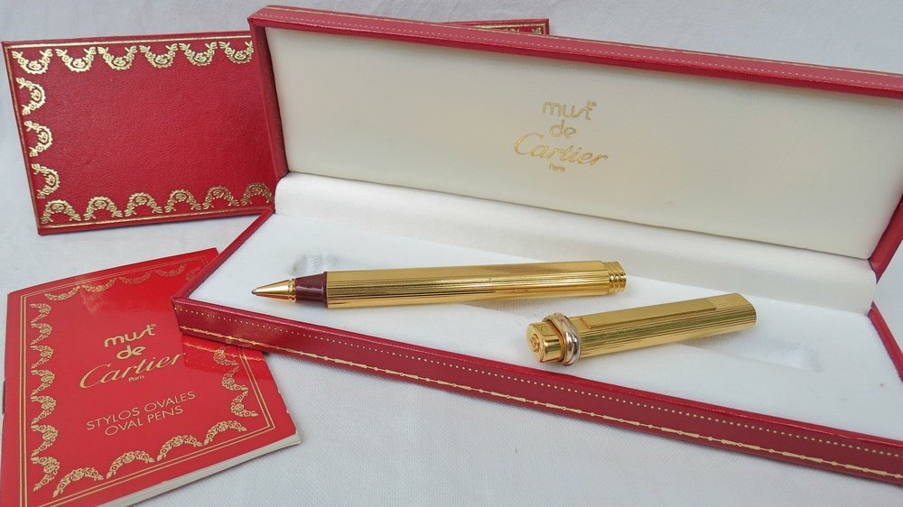 A Must de Cartier ballpoint pen, with a: A Must de Cartier ballpoint pen, with a reeded gold plated body and twisted bi-metal band to cap, within Cartier case, together with guarantee card and holder.