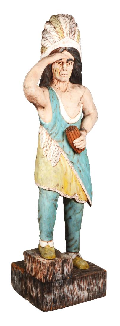 Wooden Cigar Store Indian.: Carved wood statue of a Native American Indian Chief holding a bundle of cigars in one hand while he uses the other to shade his eyes. Painted in pastel hues, displaying a weathered appearance with cr
