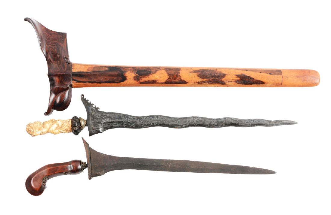 Lot of 2: Indonesian Kris.: Comprising: A) Serpentine Damascus steel blade with integral guard with decorated edge on one side. Figural Indonesian handle of bone horn or ebony base and silver ferrule. Carved wooden scabbard feat