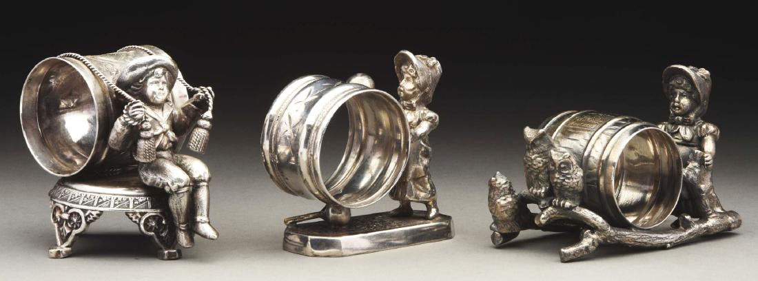 Lot of 3: Kate Greenaway-type Napkin Rings.: The first is a Kate Greenaway-type girl with drumstick stands by raised, rectangular base by pedestaled napkin holder. By Simpson, Hall, Miller & Co. Silver and condition are very good. The second is