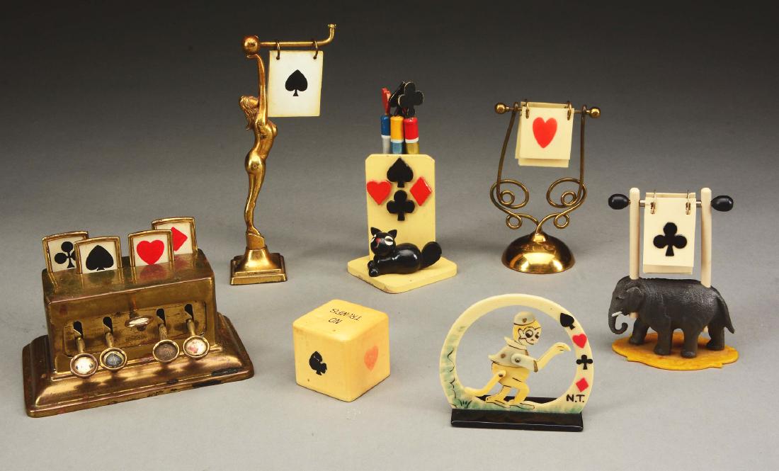Lot Of 7: Trump Markers for Cards.: Early 20th century. The first in the form of a brass cash register, a carved wood pencil holder, a celluloid Monkey in an arch, a scrolling wirework, cast brass nude, elephant and the last is in the s