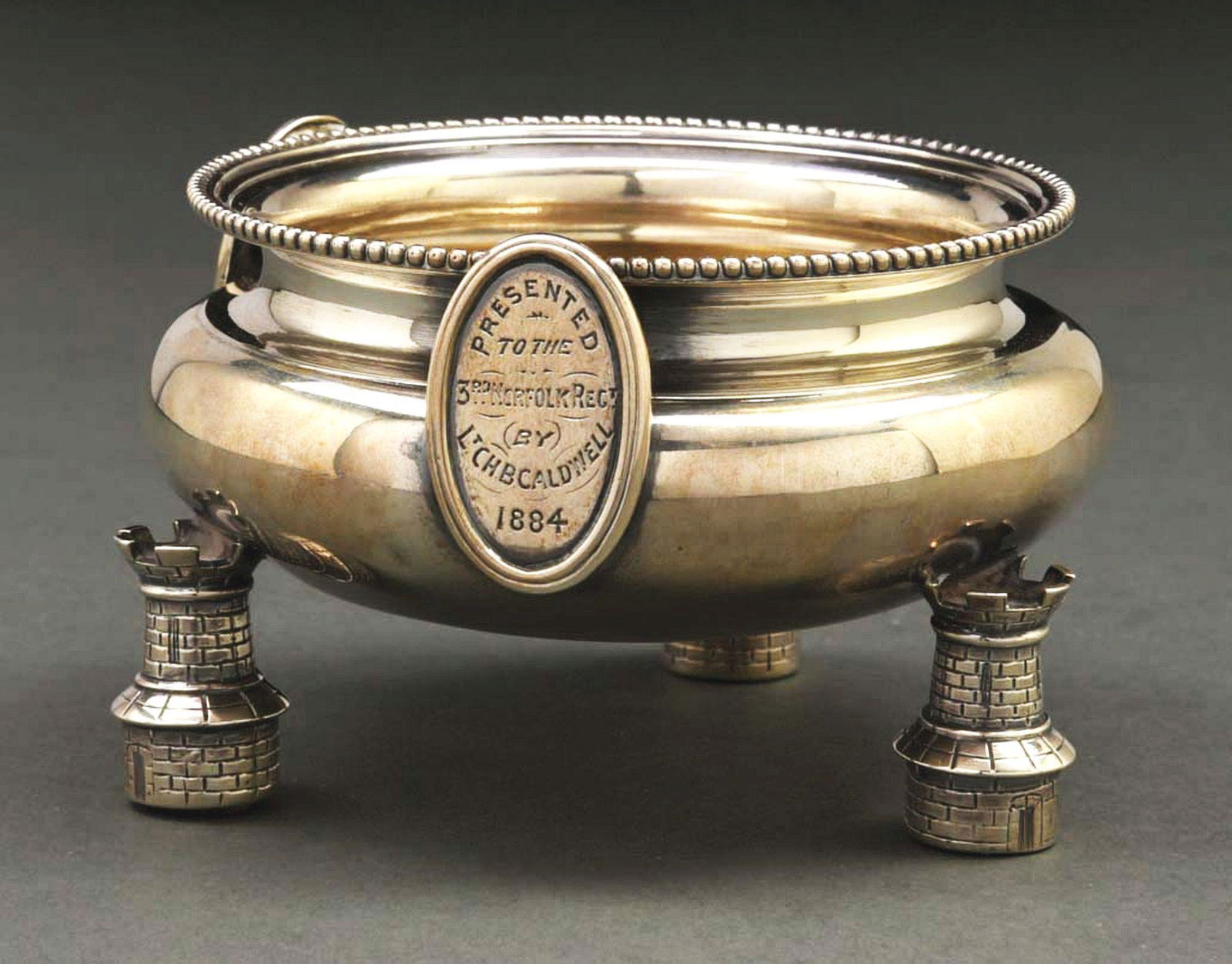 AN ENGLISH SILVER REGIMENTAL SALT CELLAR.: George Fox, London 1883, with a low circular body with a beaded rim, on three cast feet in the form of armorial towers, the sides applied with two oval plaques, one engraved with a figure of Britannia