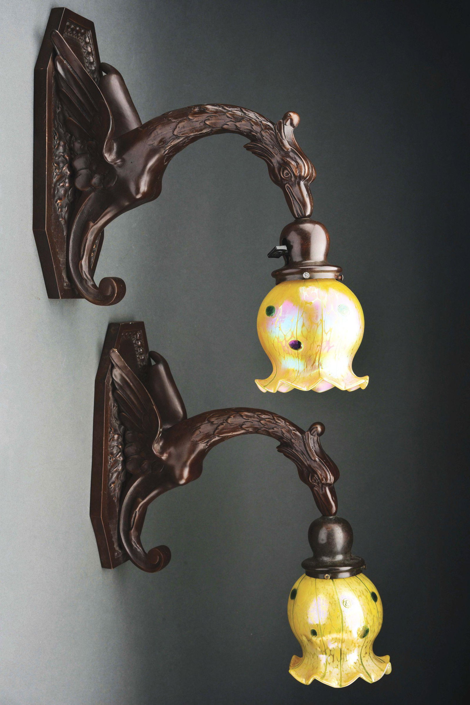ART NOUVEAU BRONZE GRIFFIN SCONCES.: A pair of patinated bronze Art Nouveau sconces in the form of a griffin, each decorated with matching Austrian art glass Loetz shades, decorated with iridescent green circles and lines against an irid