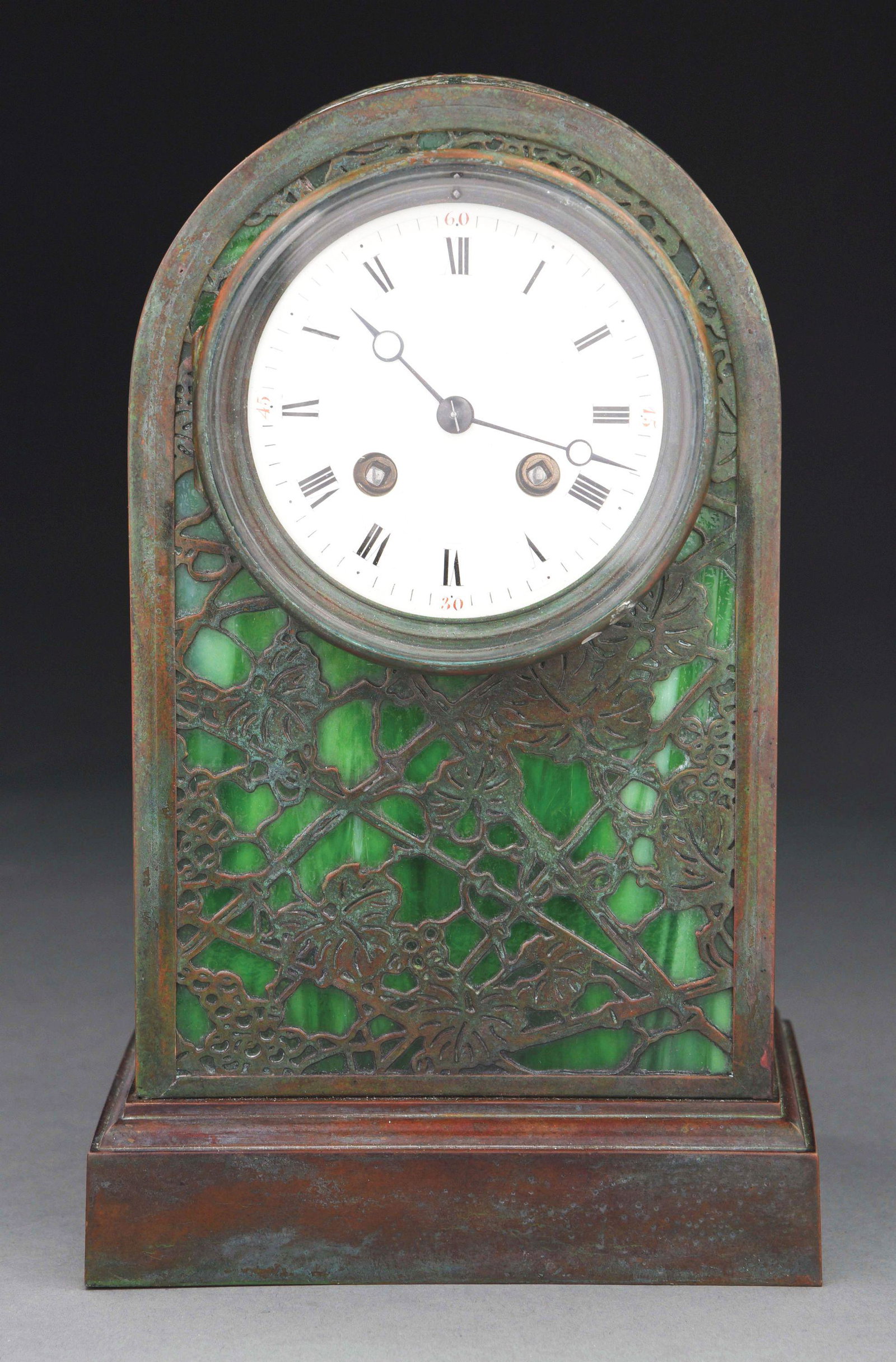 TIFFANY STUDIOS GRAPEVINE CLOCK.: A beautiful clock by Tiffany Studios. A round clock face is set in a frame of green slag glass with patinated overlay in classic grapevine pattern. The glass and clock are mounted on a patinated metal