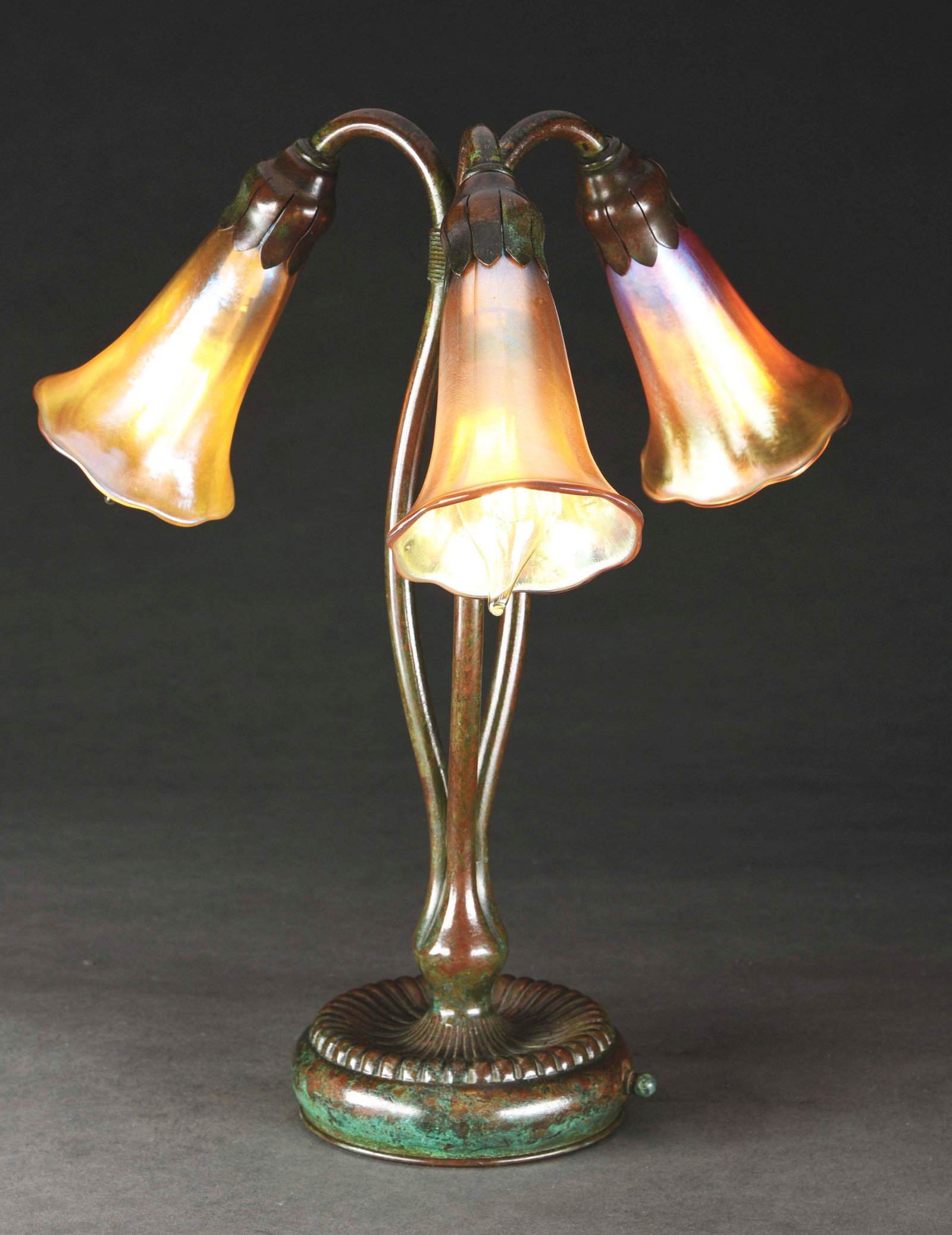TIFFANY STUDIOS LILY DESK LAMP.: Tiffany Studios patinated bronze three-light shower lily desk lamp decorated with three iridescent gold art glass lily shades set within a Tiffany Studios patinated bronze three-arm shower lily base. 