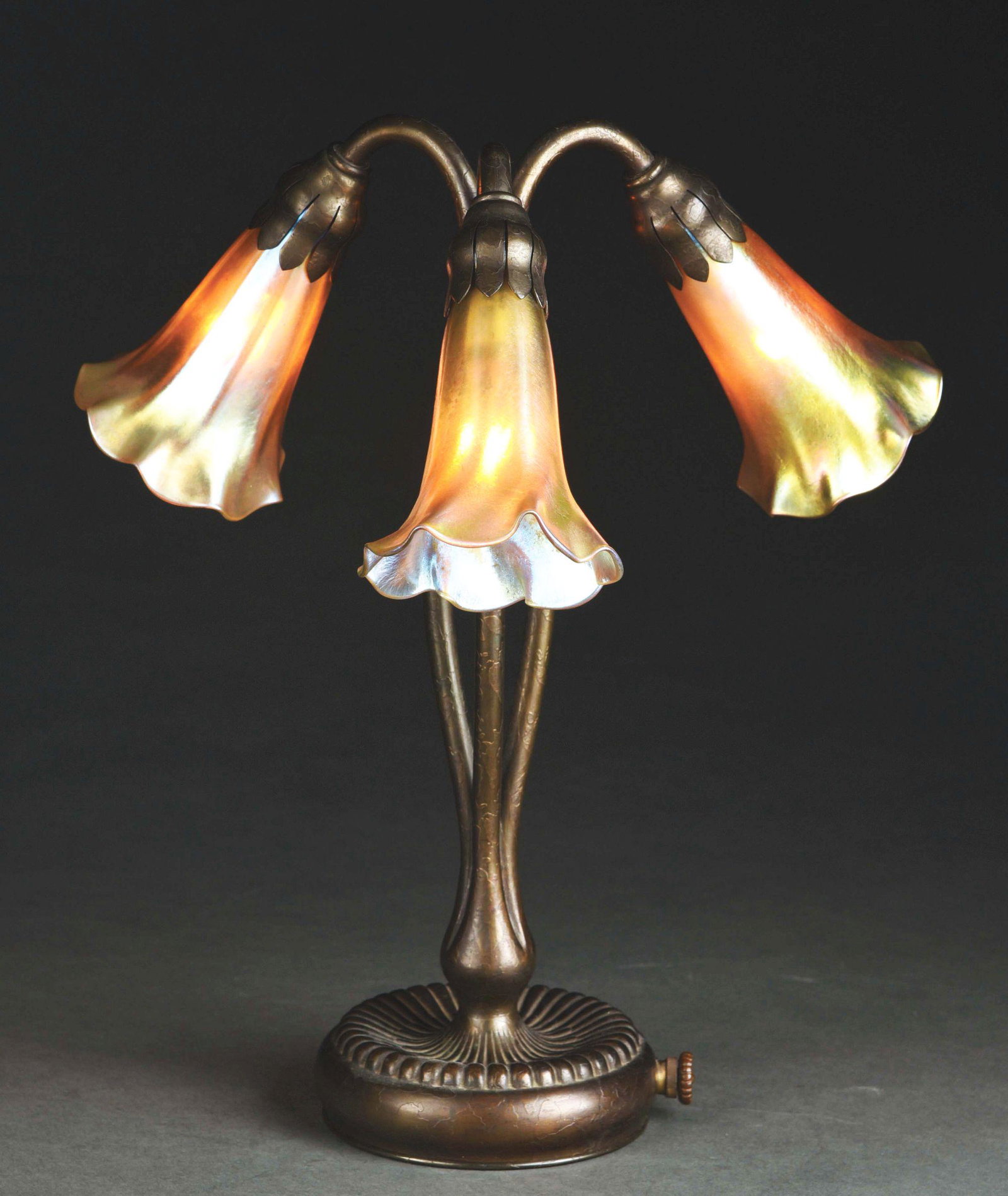 TIFFANY STUDIOS THREE-LIGHT LILY LAMP.: A favrile three-light lily lamp with bronze base by Tiffany Studios. The lamp features three favrile glass gold shades. The shades are gold with subtle iridescence throughout. The shades are ruffled a