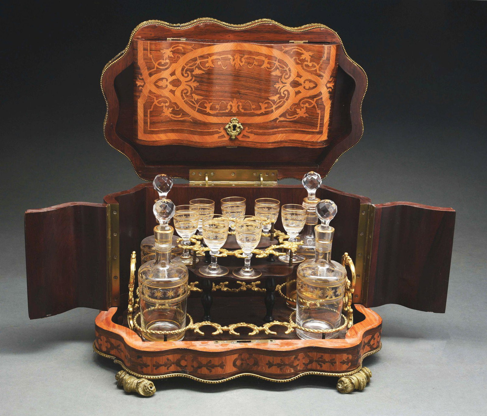 ANTIQUE TANTALUS WITH INLIAD WOOD DECANTER LIQUER: Antique tantalus decanter liqueur opens to display four crystal decanters and six stemmed glasses with repairs. Exquisite workmanship to inlaid cabinet with brass overlay, c. 1880. One large chip or c