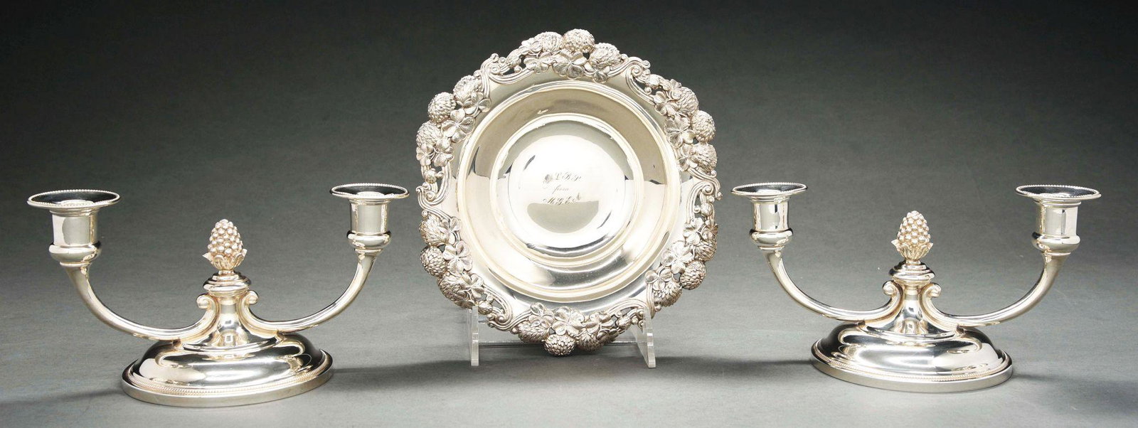 A TIFFANY STERLING BOWL AND A PAIR OF LOW CANDELABRA.: The circular bowl with a shaped edge applied with strawberries around a plain center with initials; the low two-light candelabra with broad domed bases and plain sockets centering pomegranate finials.