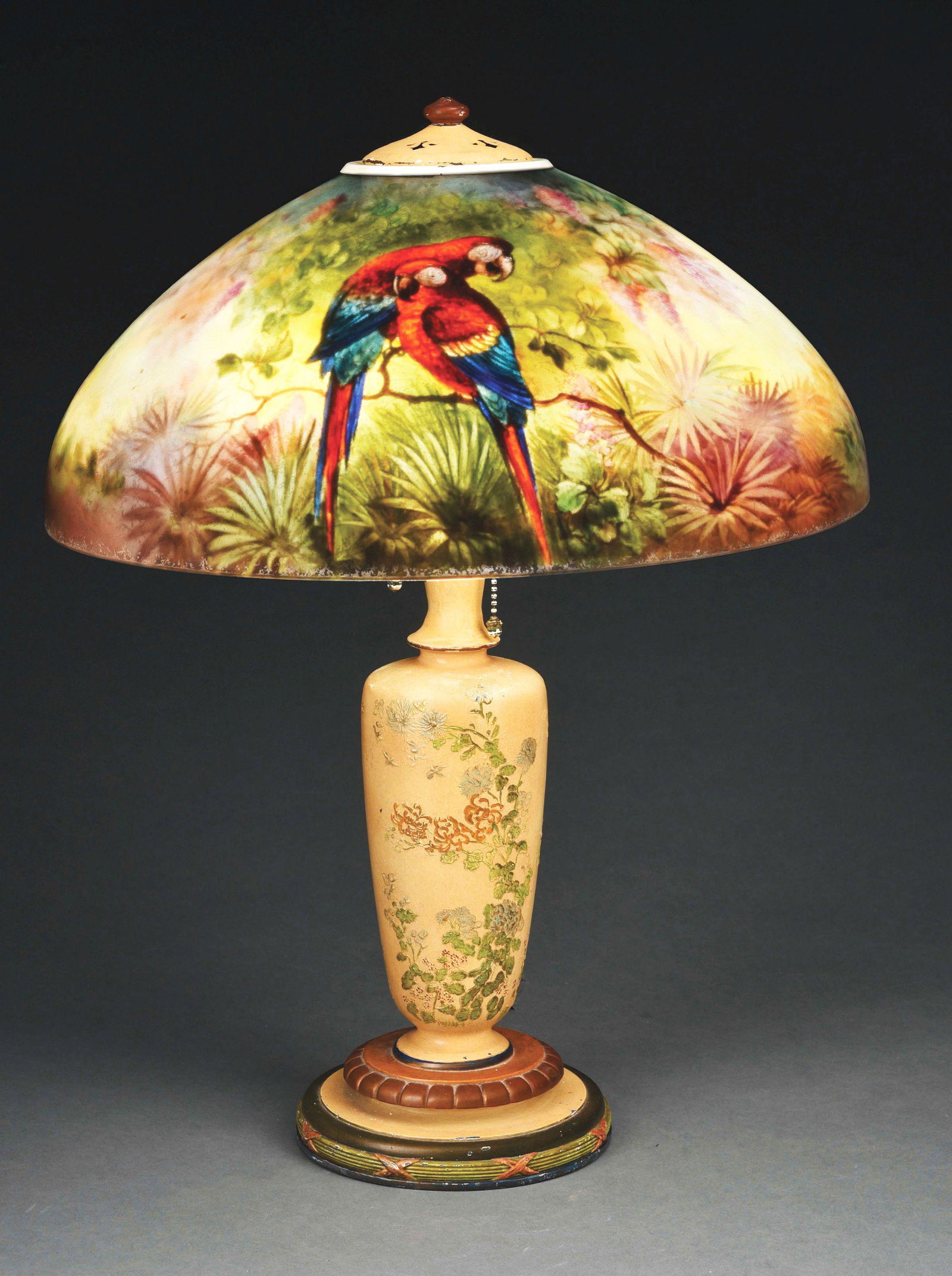 HANDEL REVERSE PAINTED PARROT TABLE LAMP.: A beautiful reverse painted table lamp with original base by Handel. The 18” reverse painted shade features a jungle scene with a pair of crimson and blue parrots resting on a branch, and a third pa