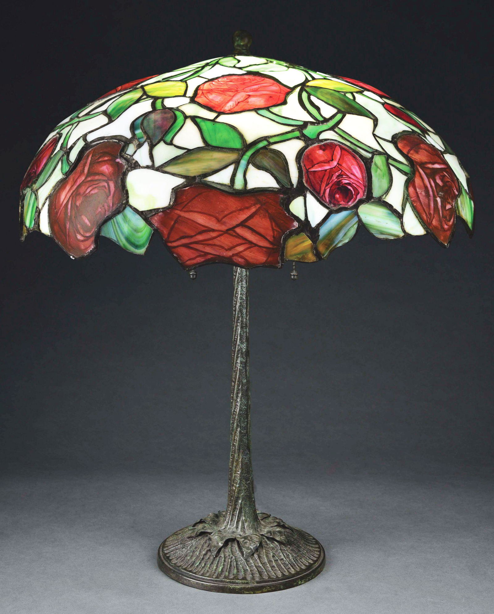 JOHN MORGAN AND SONS ROSE LEADED GLASS TABLE LAMP.: A beautiful Rose leaded glass lamp by Morgan. The shade features painted red roses with green leaves, set against a background of green and white glass. The shade measures approximately 20” in diame