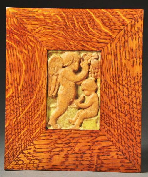 Grueby 2-Color Faience Tile.: Depicts cherubs and grapes in a contemporary oak frame. One small corner chip is visible. Size: Framed: 11 - 3/4" x 9 - 3/4".