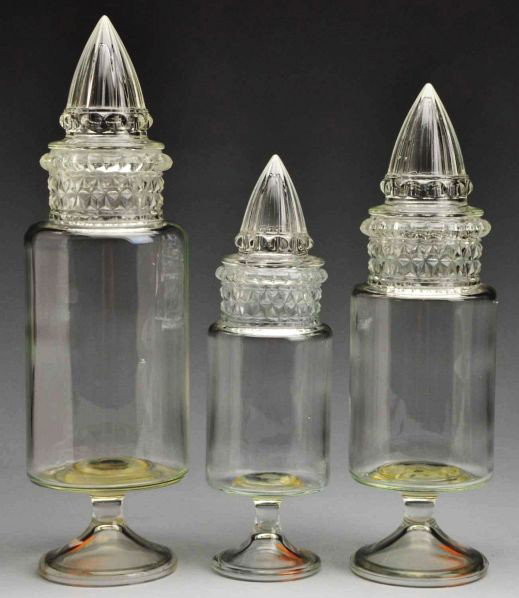 Lot of 3: Heck Spired Cylinder Apothecary Jars.: Circa early to mid-20th century. Set of three matching clear glass Heck spired cylinder jars. Pedestals attached to jar bottoms. Hard-to-find set of three different sizes of this rare style. Condition