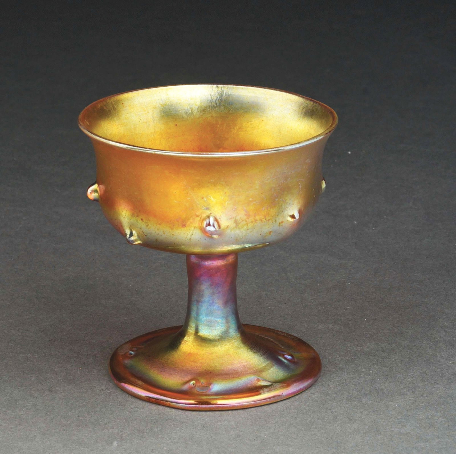 TIFFANY STUDIOS FAVRILE GLASS CUP.: A beautiful glass cup by Tiffany Studios. The 3 - 1/2" tall cup is composed of gold Favrile glass with iridescent tones in purples and pinks. It is embellished with small, protruding bumps on both the