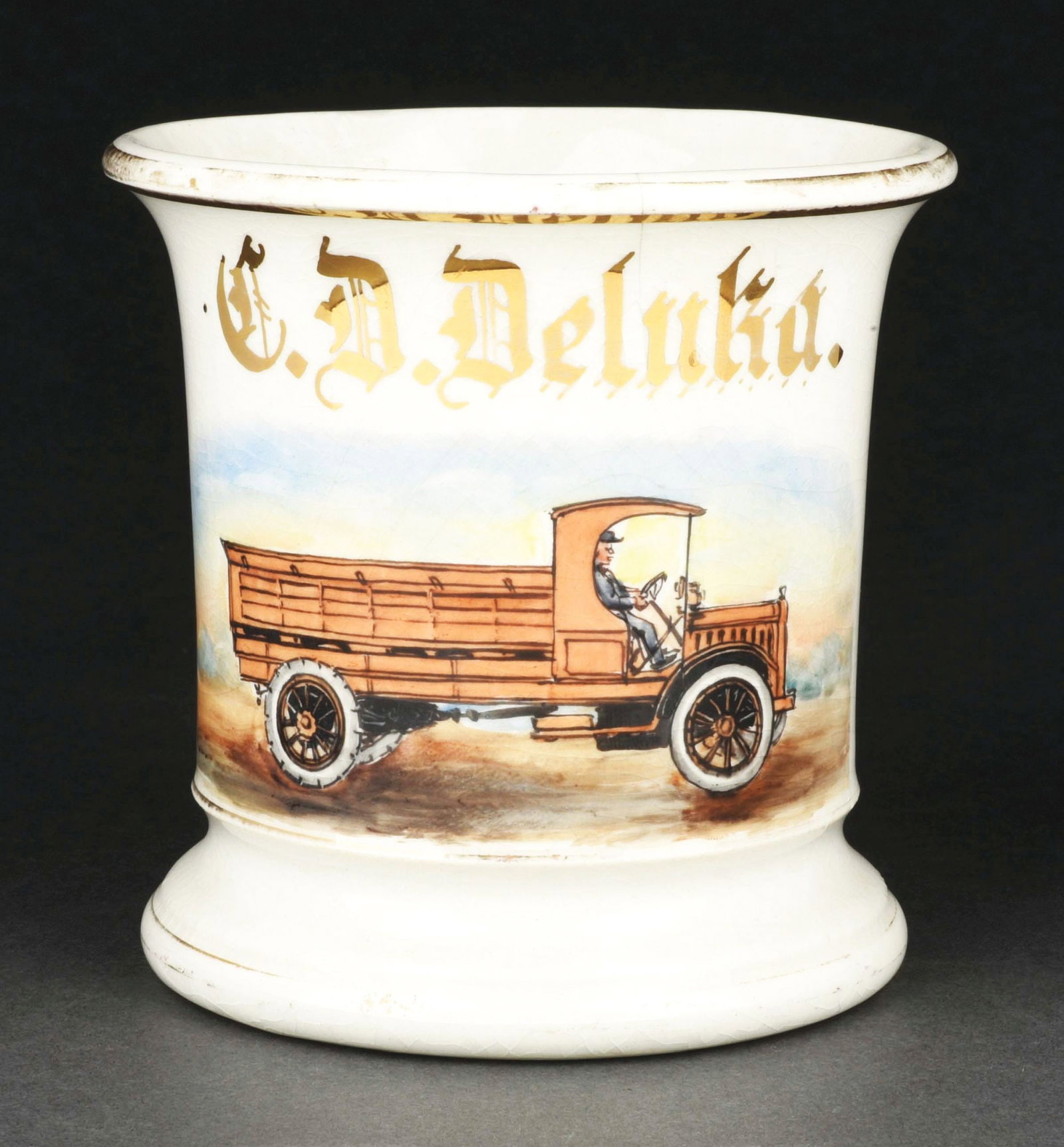 TRUCK OPERATOR SHAVING MUG: Has the name "C.D. Deluka" in gold gilt. Mug is stamped "Potters Cooperative Company" under base. Detailed image of men driving delivery truck. Minor crazing throughout. Condition: (Excellent) Item Di