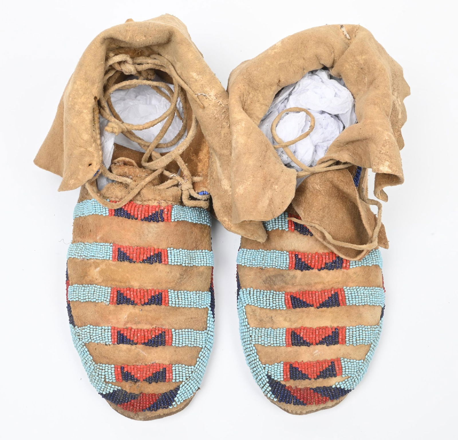 BLUE AND RED BEADED CROW MOCCASINS: Classic pair with Crow men's moccasins with powder blue, faceted red, and cobalt blue striped panels. Ankle high buckskin for use in winter months. 10 1/4" Length. CONDITION: Very good, supple. Dimens