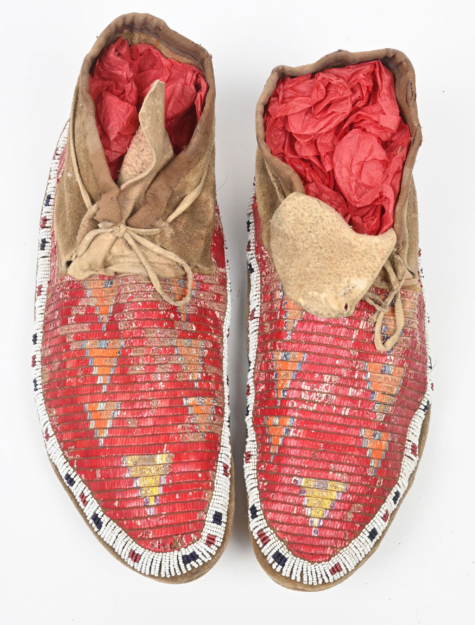 RED BEADED MOCCASINS WITH GEOMETRIC SHAPES: Plains moccasins with fully quilled vamps on Indian tanned buckskin of geometric designs on a red background. Edge beading of white heart rose, dark blue and white. CONDITION: Good with some light bug