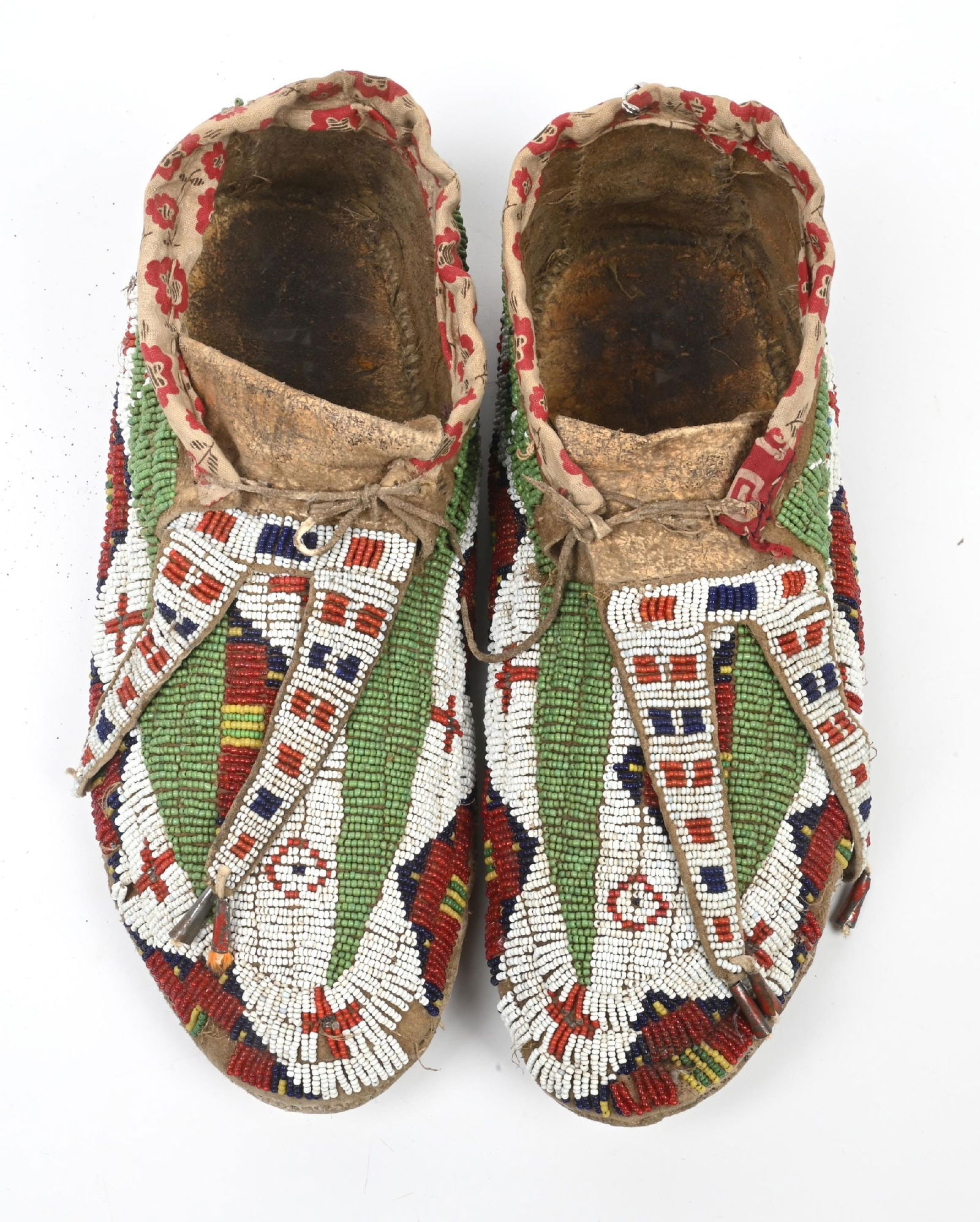 GREEN & WHITE BEADED ARAPAHO MOCCASINS: Sinew sewn geometric designs of greasy yellow, cobalt blue, red, and white patterns on a dusty green field. Rawhide soles with calico lined cuffs. 11" Length. CONDITION: Very good with some light bead