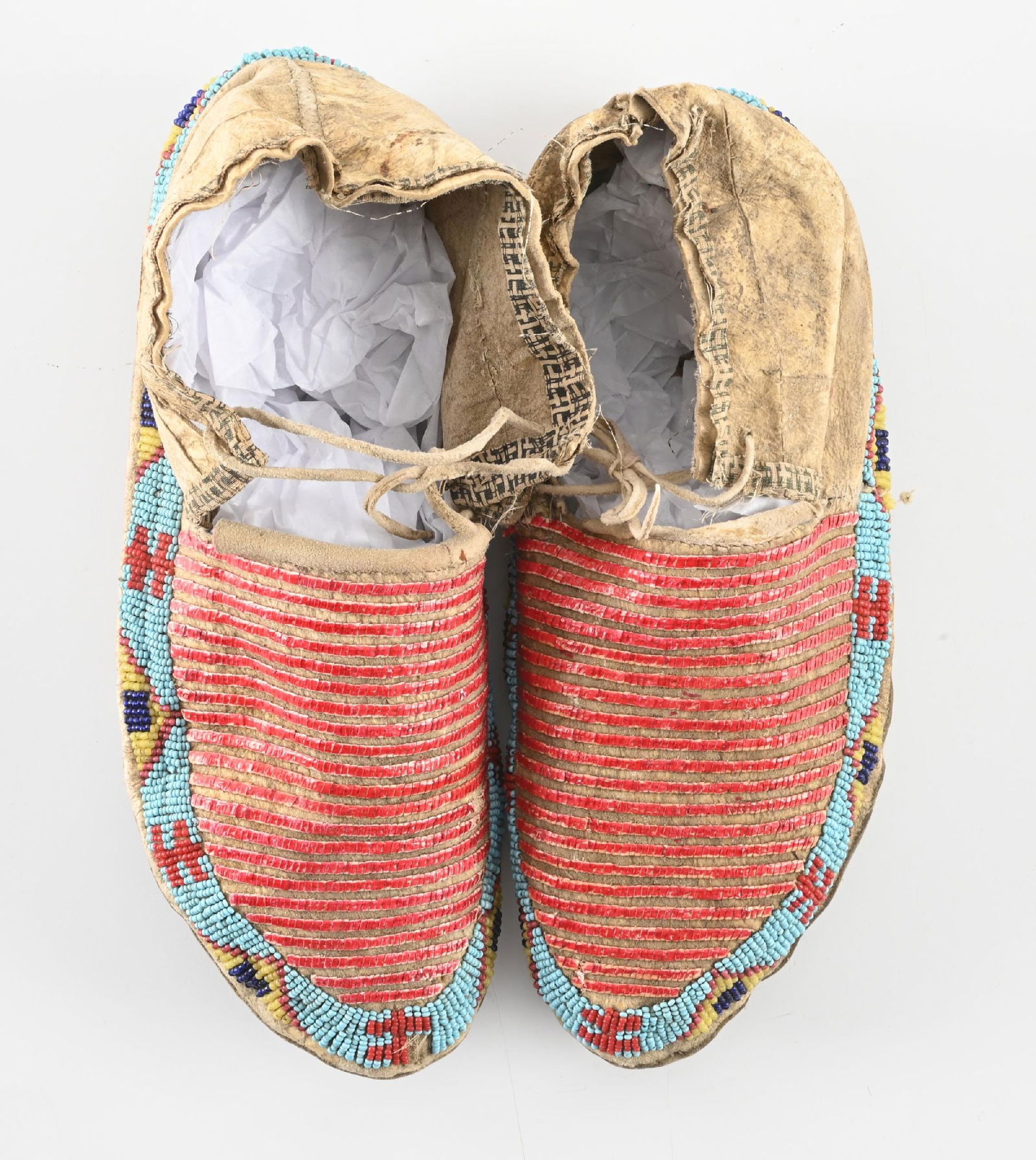 SIOUX MOCCASINS: Lakota moccasins finished in bands of red porcupine quill with edge beading in greasy yellow, red, white heart rose, powder blue and translucent blue. Soles are rawhide with calico cloth edged uppers.