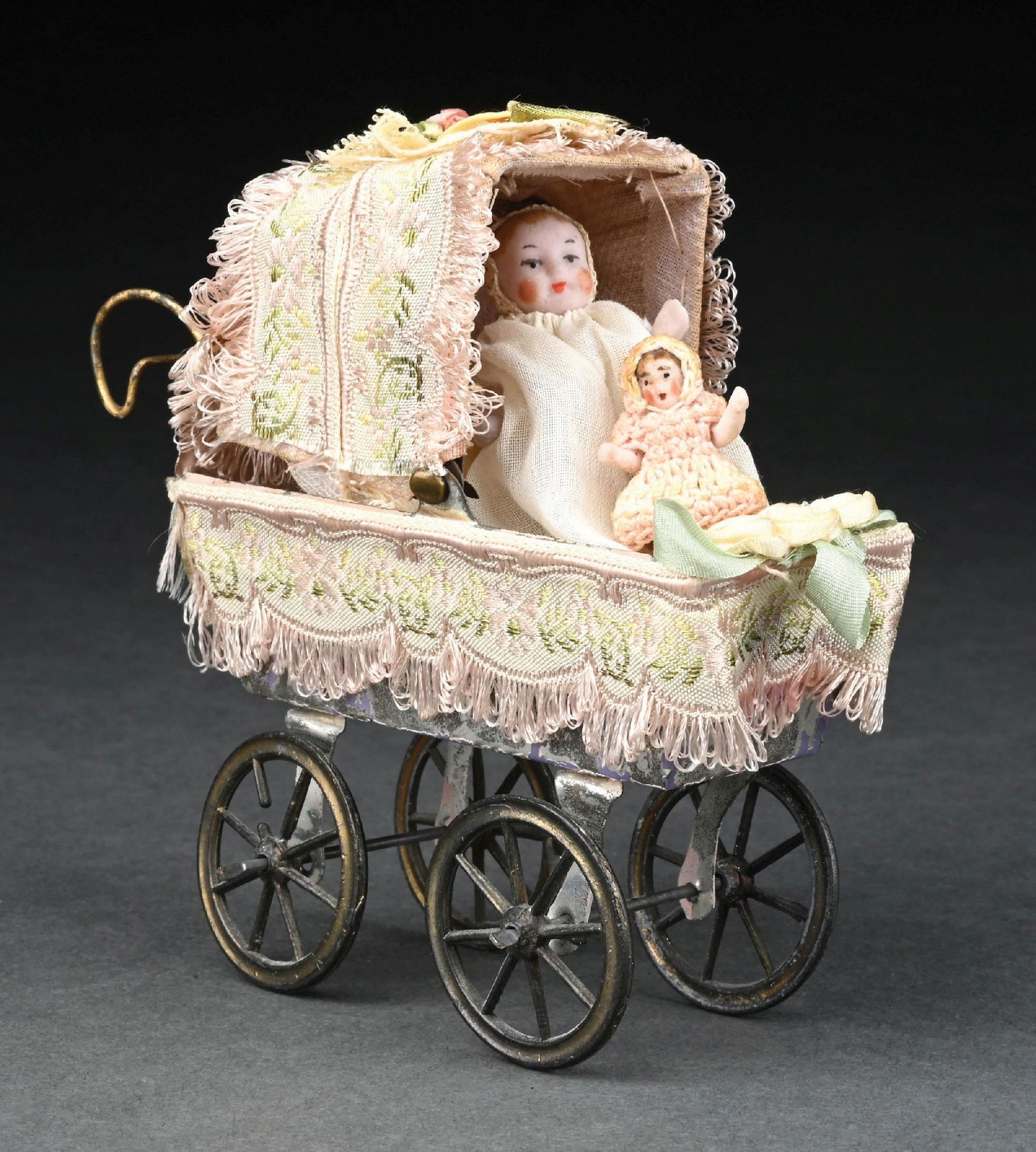 UNUSUAL GERMAN TIN PLATE BABY CARRIAGE W/ SPOKED WHEELS: The spoke wheels have been recovered at some point in a fancy brocade material with pink lace and silk floral adornments on the top. Inside are two very small Bisque babies, one stands alone and the o
