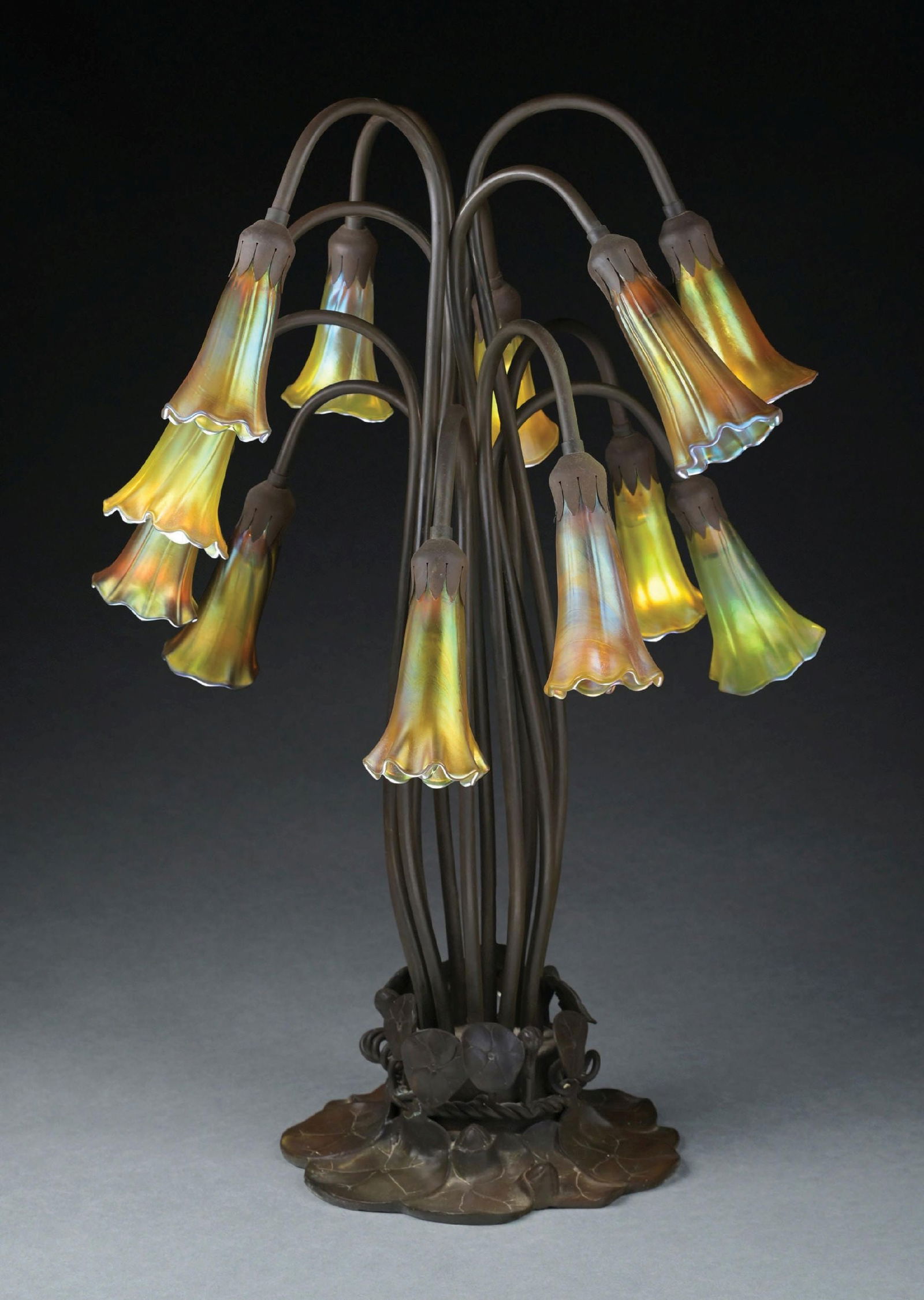 TIFFANY-STYLE 12-LIGHT LILY LAMP: A lovely Tiffany-style 12-light art glass and bronze lily lamp. The lamp has 12 gold art glass lily shades. The shades have lovely iridescence and prominent ribbing on most. The shades sit in a lily p