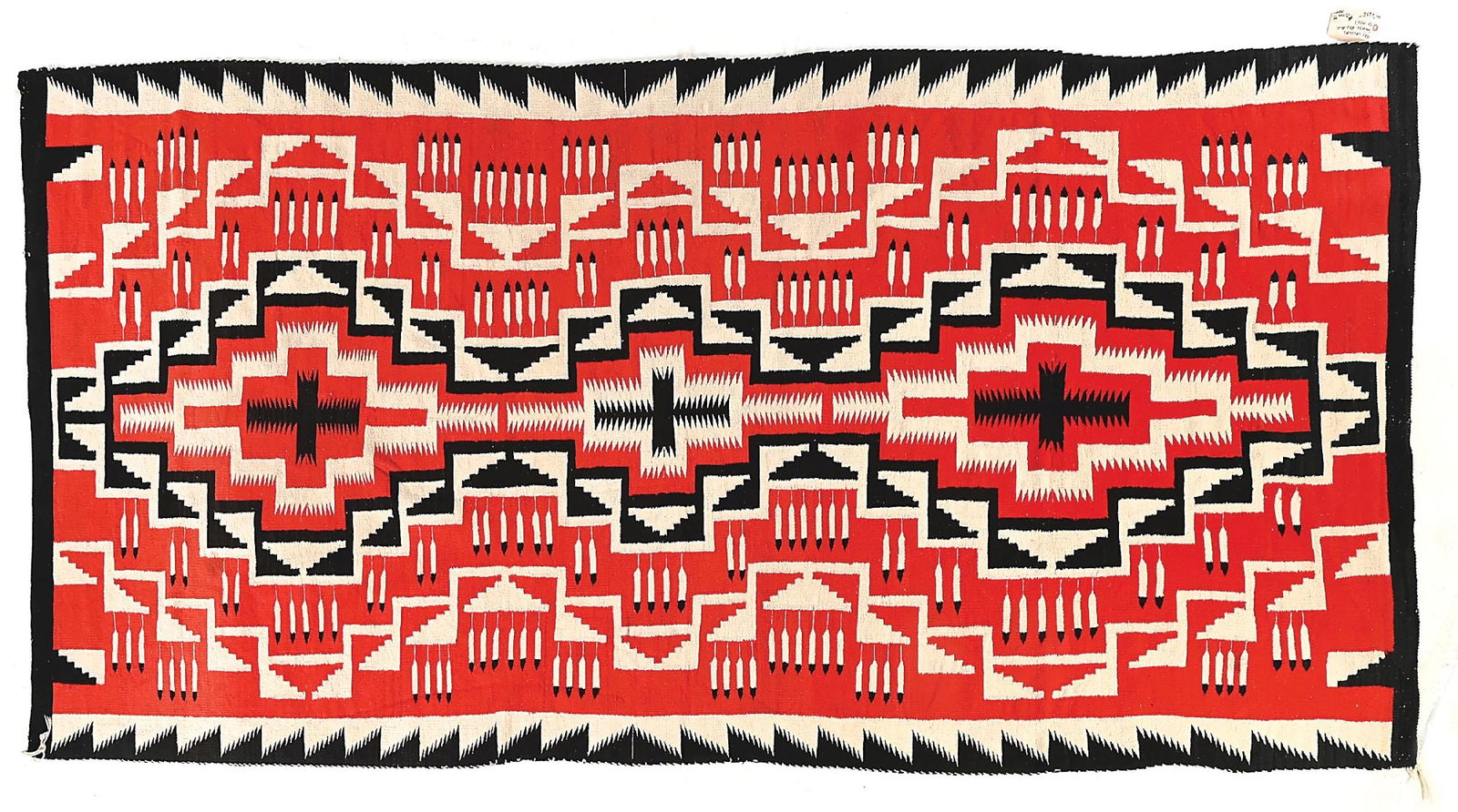 NAVAJO (DINÉ) LARGE RUG GANADO: Contemporary. A central vertical column of three-step diamonds, each containing serrated cross patterns, flanked on each side by eagle feather motifs and stepped triangles, encased by side serrated bo