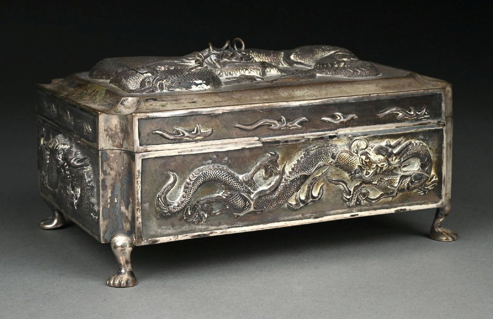A CHINESE SILVER BOX: Retailer’s mark of Zeewo, rectangular with incurved corners and a with a hinged lid chased with a dragon, the sides with smaller dragons, and on four animal paw feet. Length: 8-1/2". Weight: